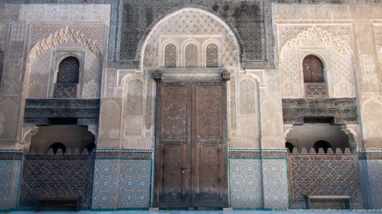 Tall carved double wooden door framed by pale plaster ornament, dark wood screens, and blue-green geometric tilework.