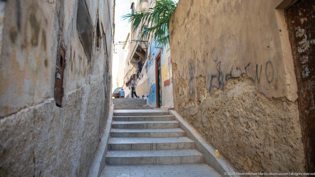 Stone steps rising through a narrow medina passage between tall weathered plaster walls.