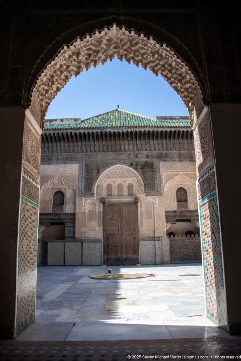 Sunlit decorated courtyard façade seen through a shaded horseshoe arch with mosaic-tiled lower walls.