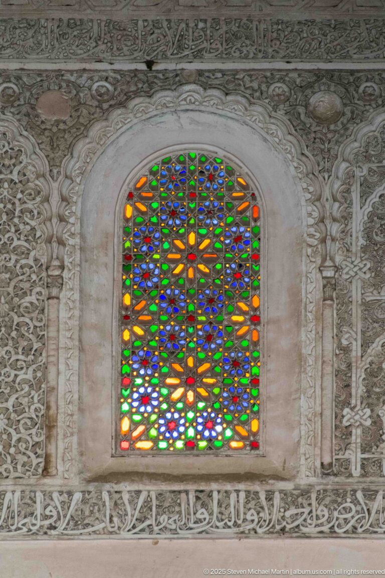 Arched window filled with multicolored geometric glass and surrounded by densely carved pale stucco.