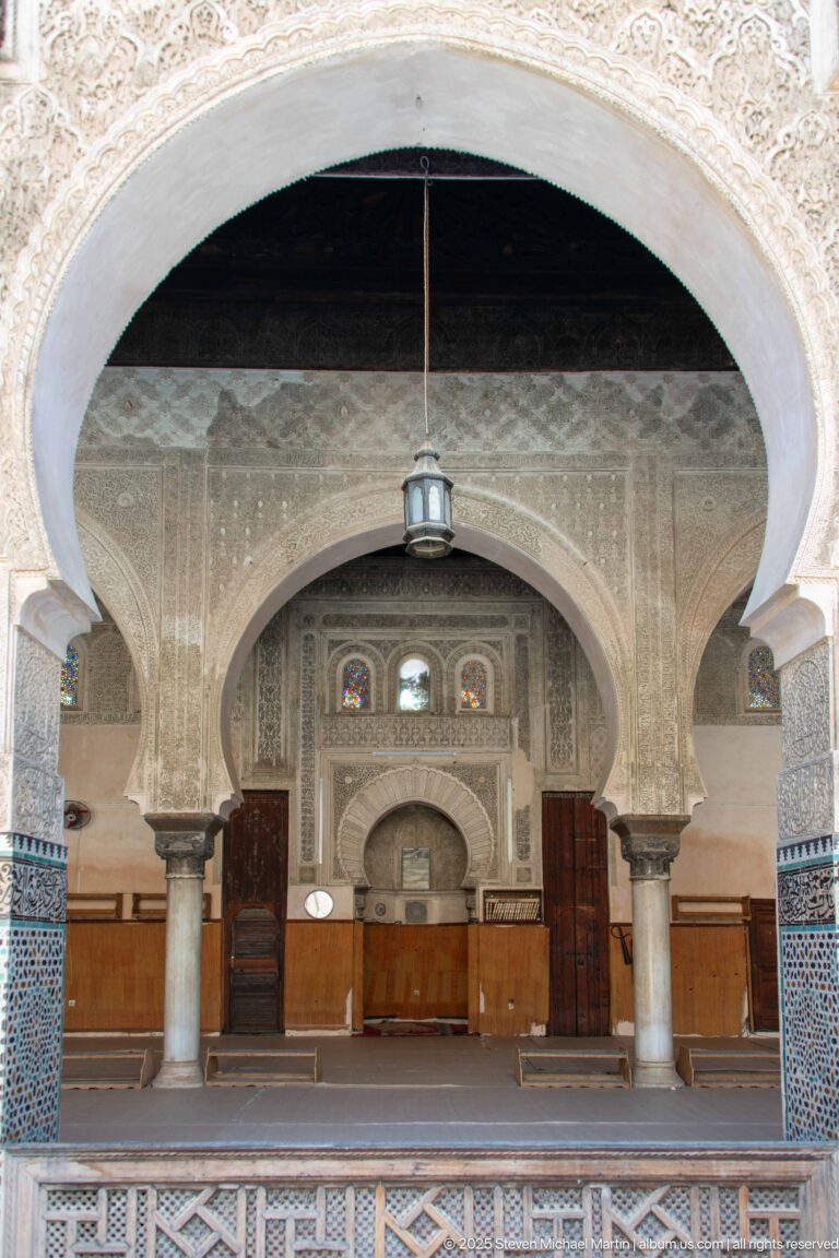 Broad horseshoe arch opening to an arcaded interior with columns, carved walls, tilework, woodwork, and a hanging lantern.