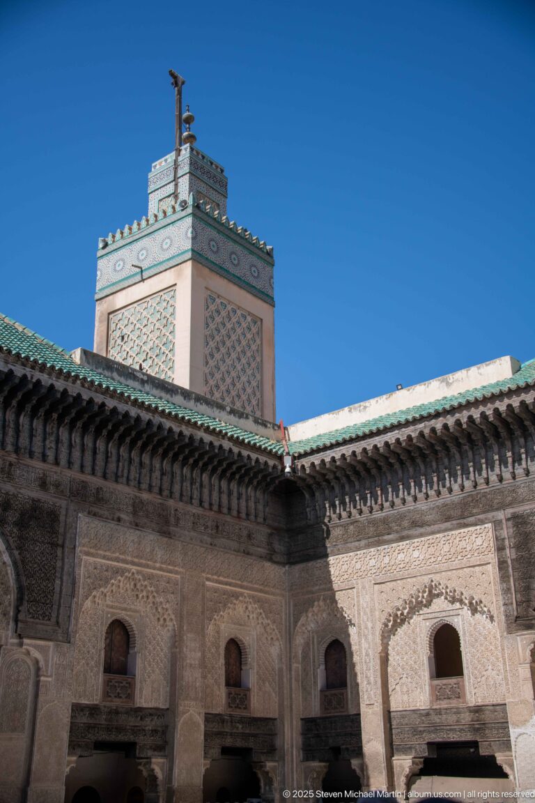 Square decorated minaret rising above carved courtyard walls and green-tiled eaves against blue sky.
