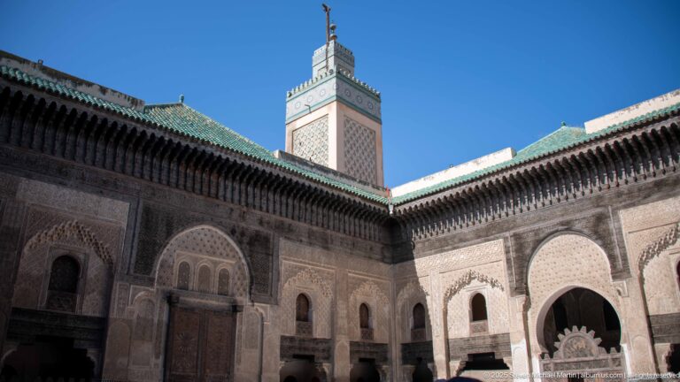 Two decorated courtyard façades meeting below a square minaret, with carved plaster, doors, tilework, and timber eaves.