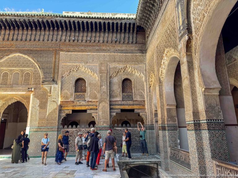 Visitors standing beside a decorated courtyard wall with carved plaster, mosaic tilework, wooden eaves, and a large arch.