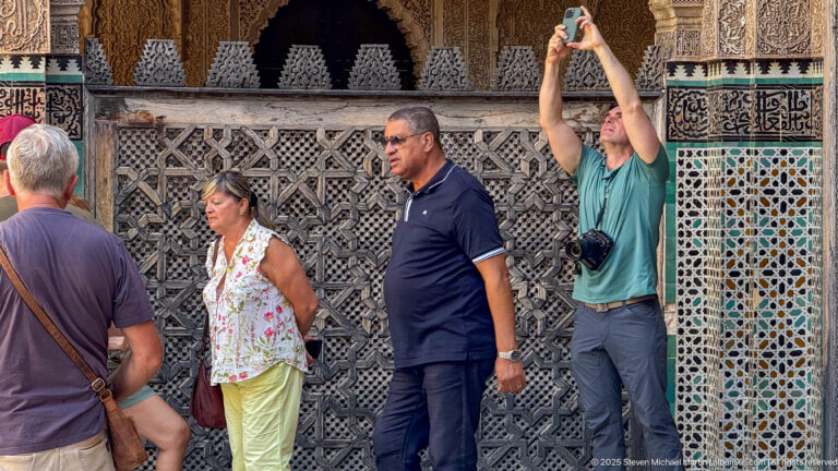 Visitors beside a dark carved wooden screen and multicolored zellij panel; one person holds a camera overhead.