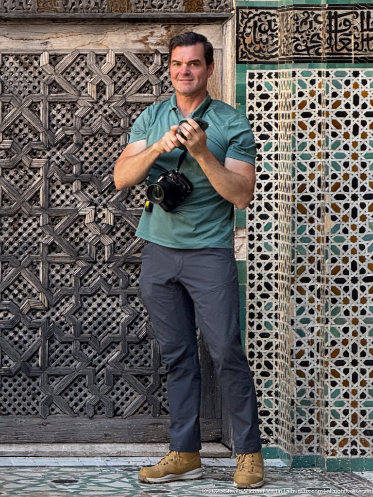 Man with camera standing beside a large carved wooden screen and a vertical geometric zellij panel.
