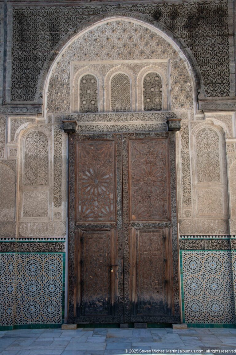 Symmetrical double wooden doorway framed by carved stucco arches and blue-green geometric tilework.