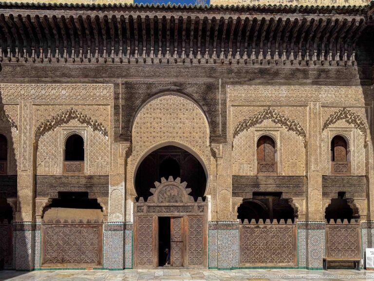 Frontal decorated courtyard façade with central wooden door, large horseshoe arch, smaller windows, and mosaic tilework.