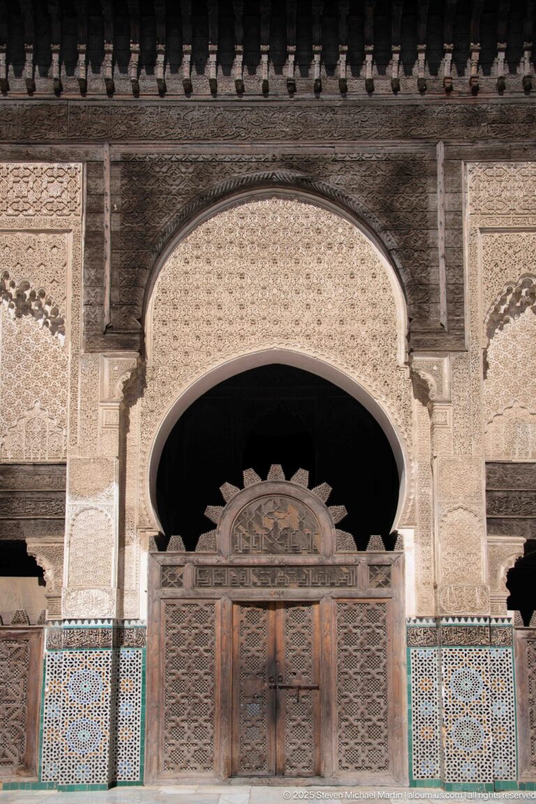 Wooden double doors beneath a scalloped horseshoe arch, surrounded by carved plaster and blue-green zellij.