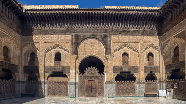 Wide frontal view of a symmetrical courtyard wall with central doorway, arched windows, carved plaster, tilework, and timber eaves.