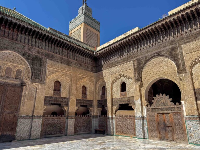 Oblique courtyard view with square minaret, decorated walls, arched recesses, tilework, and projecting wooden eaves.