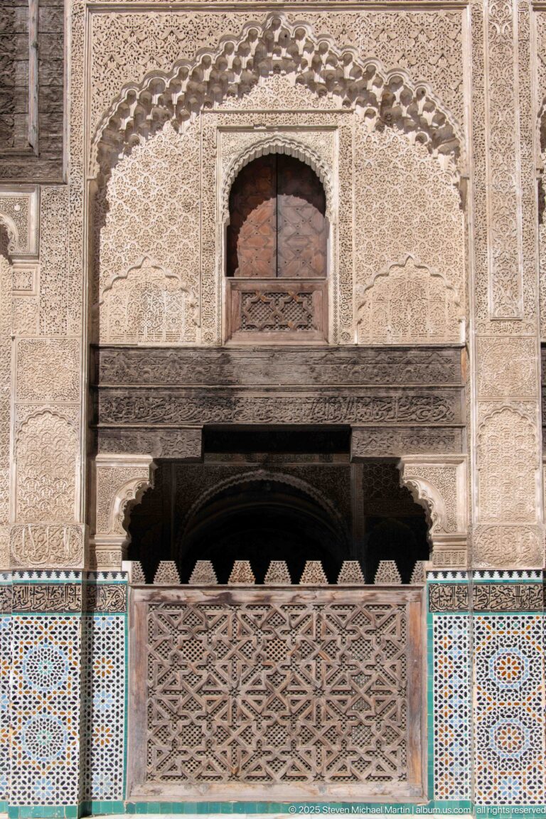 Decorated wall bay with small upper opening, carved plaster, pierced wooden balustrade, and multicolored zellij panels.