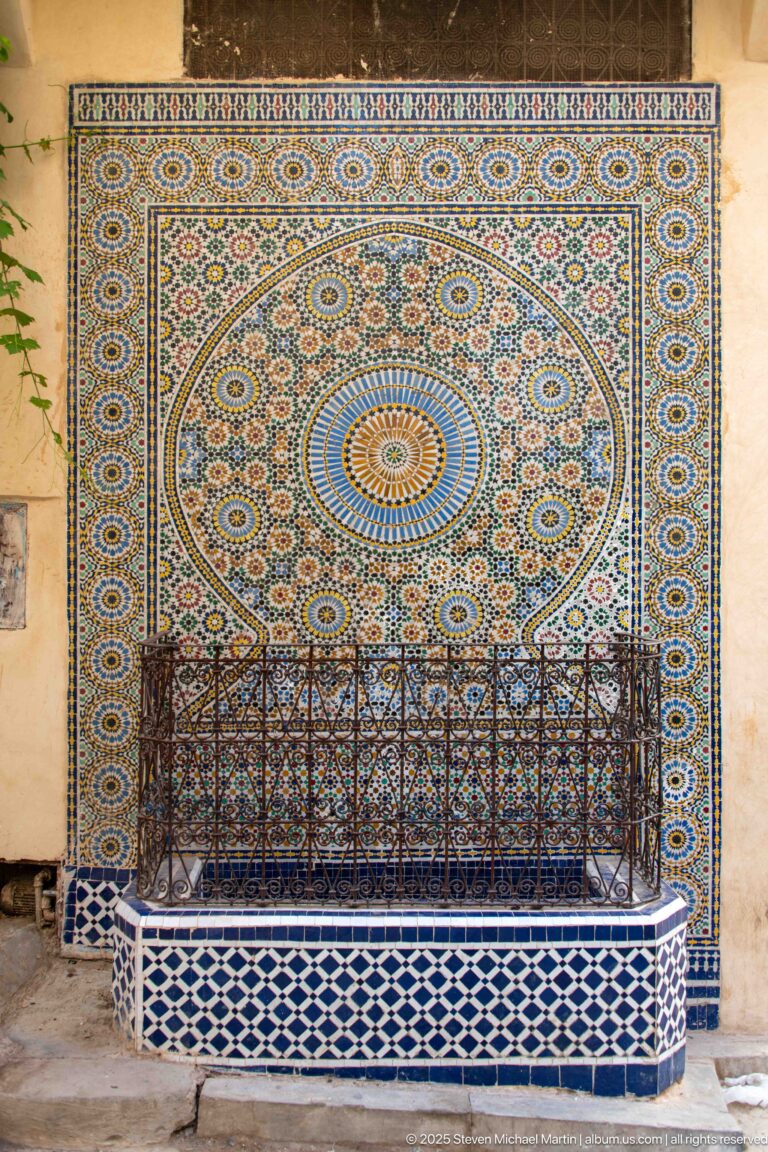 Wall fountain covered in multicolored zellij with a large circular rosette and iron grille over the basin.