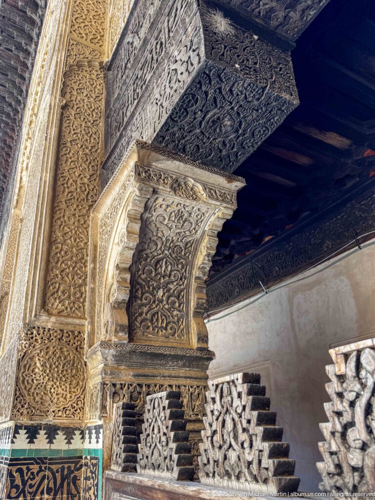 Oblique view of a richly carved stucco pier and capital beneath a dark timber ceiling, with zellij at the base.
