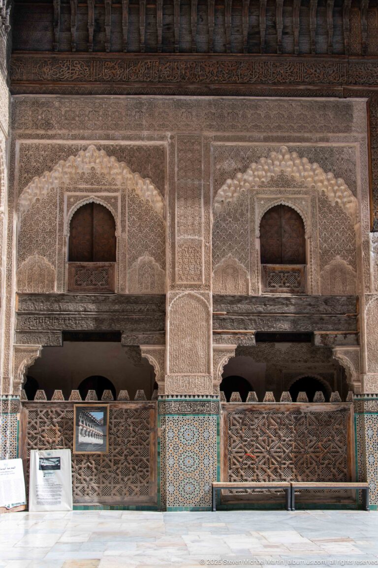 Two matching decorated wall bays with arched openings, carved plaster, wooden screens, zellij, and a bench below.
