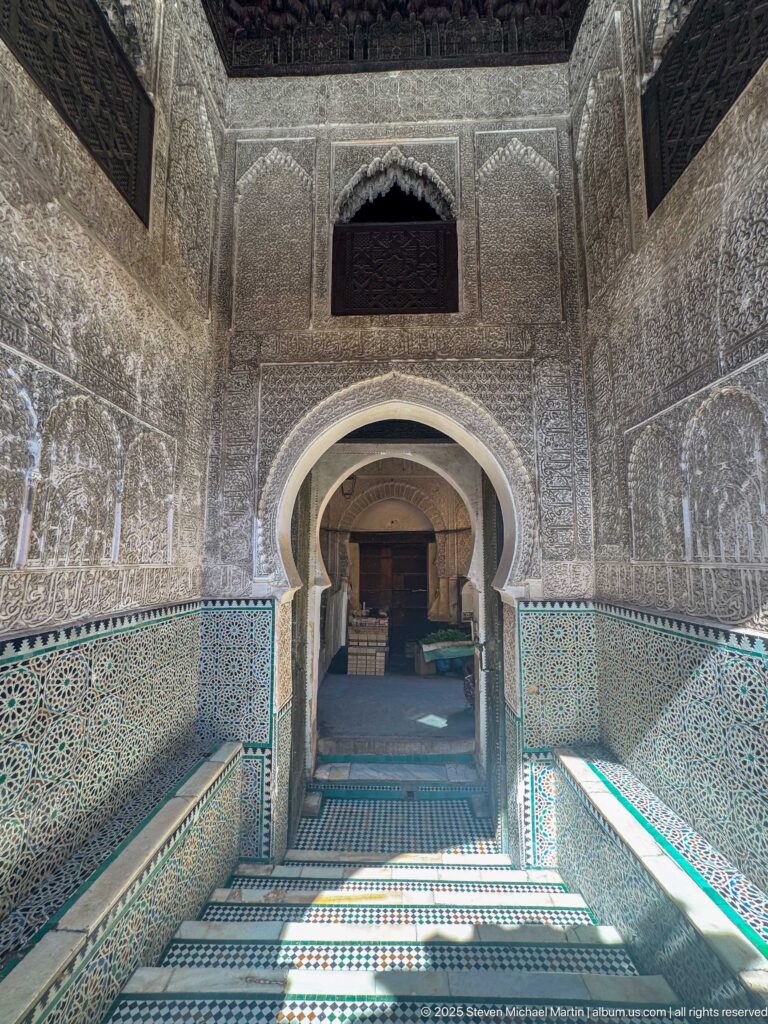 Long corridor with nested horseshoe arches, blue-green zellij walls, carved plaster, and striped sunlight on the floor.