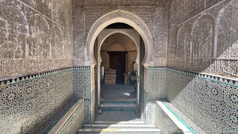 Stairway leading to a horseshoe-arched doorway between zellij lower walls and densely carved stucco surfaces.