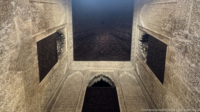 Upward view between densely carved plaster walls with dark wooden window grilles and a dark timber ceiling.