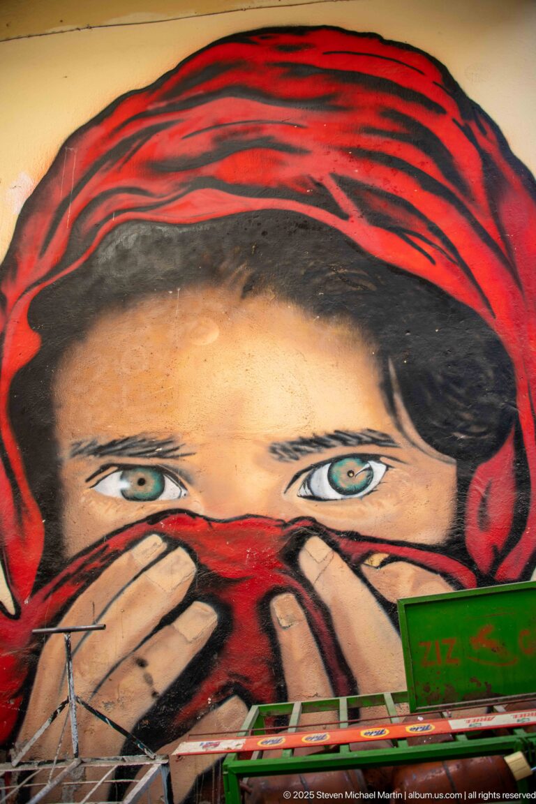 Large street mural showing a woman’s eyes above a red face covering and beneath a red-and-black head covering.