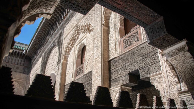 Ornately carved courtyard façade with arched openings, timber galleries, stucco decoration, and deep shadows in Fes Medina.