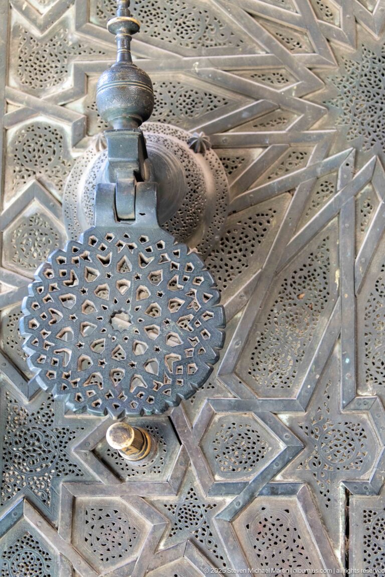 Close-up of a circular perforated metal fitting set into a silver-toned surface covered with geometric punched decoration.