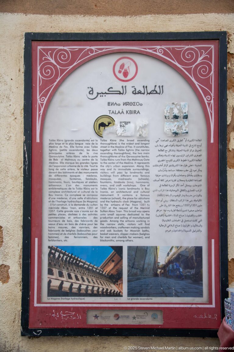 Red-bordered bilingual interpretive panel with text blocks and two small photographs mounted on a wall in Fes Medina.