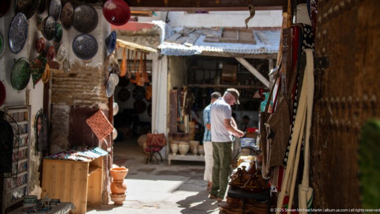 Visitor browsing pottery, textiles, and metal goods in a narrow sunlit market passage in Fes Medina.
