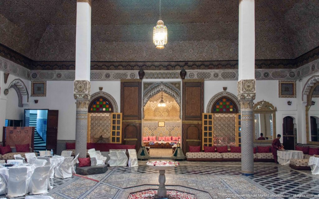 Symmetrical palace hall with central fountain, two columns, ornate salon alcove, colored-glass windows, and white-covered dining chairs.