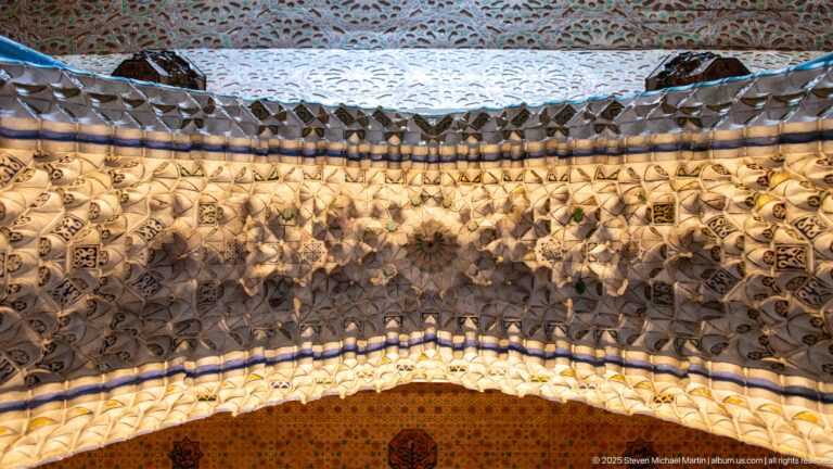 Close-up of a deep faceted muqarnas-like band over an arch, bordered by blue and painted ornament.