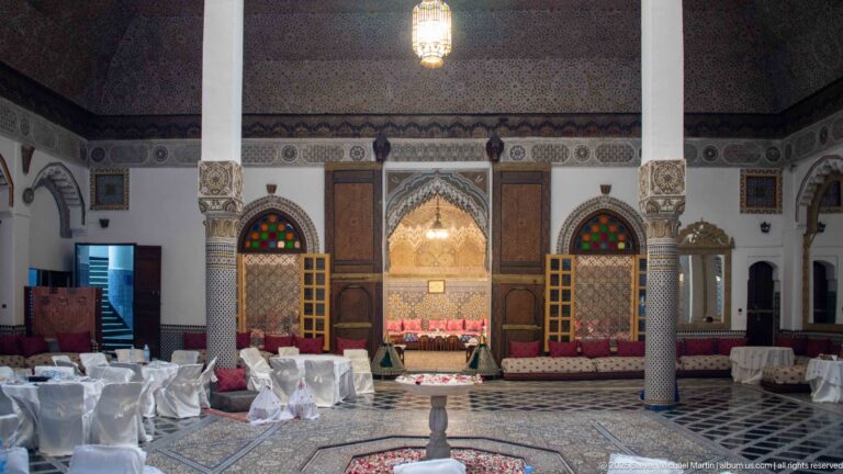 Wide columned palace hall with tiled floor, small central fountain, low seating, ornate recess, and white-covered chairs.