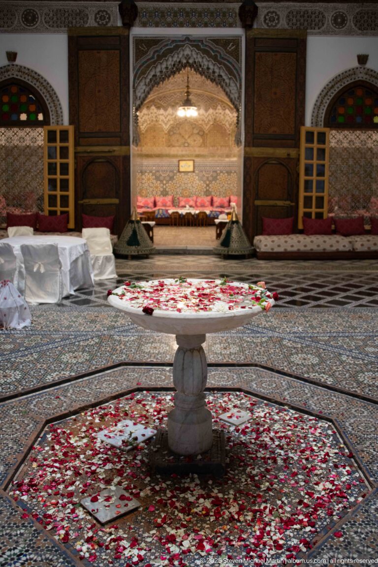 Small pedestal fountain surrounded by red and white flower petals on a geometric tiled floor, with salon seating beyond.