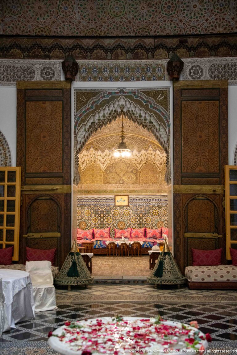 Symmetrical reception wall with carved arched alcove, zellij, bright cushions, wooden panels, and flanking seating bays.