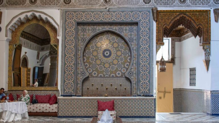 Large arched zellij fountain panel with circular mosaic rosette and lower basin, flanked by palace arches and salon seating.