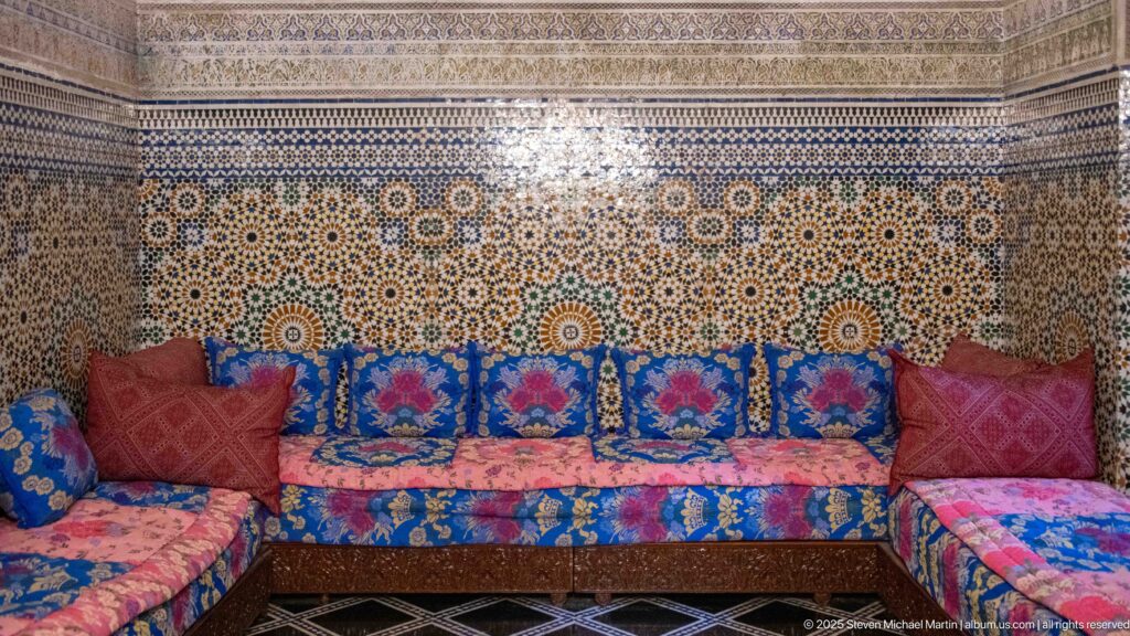 Colorful low salon seating beneath a wall of intricate geometric zellij mosaic and a carved decorative frieze.