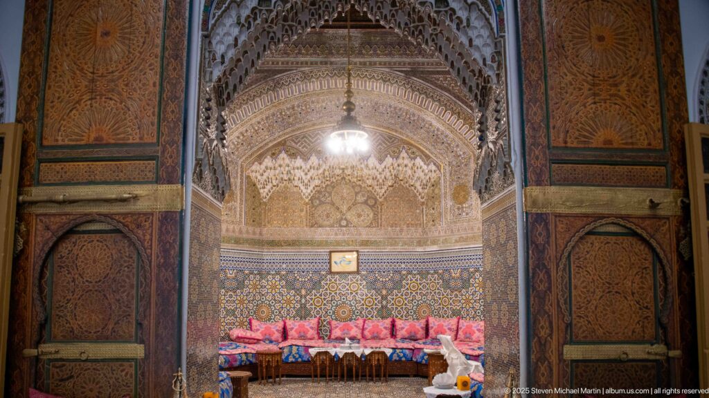 Dark carved wooden panels framing a bright salon alcove with hanging light, zellij, patterned plaster, and colorful cushions.