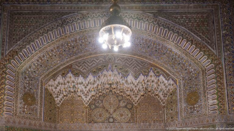 Close frontal view of a richly patterned arch with projecting white muqarnas-like carving and a hanging lamp above.