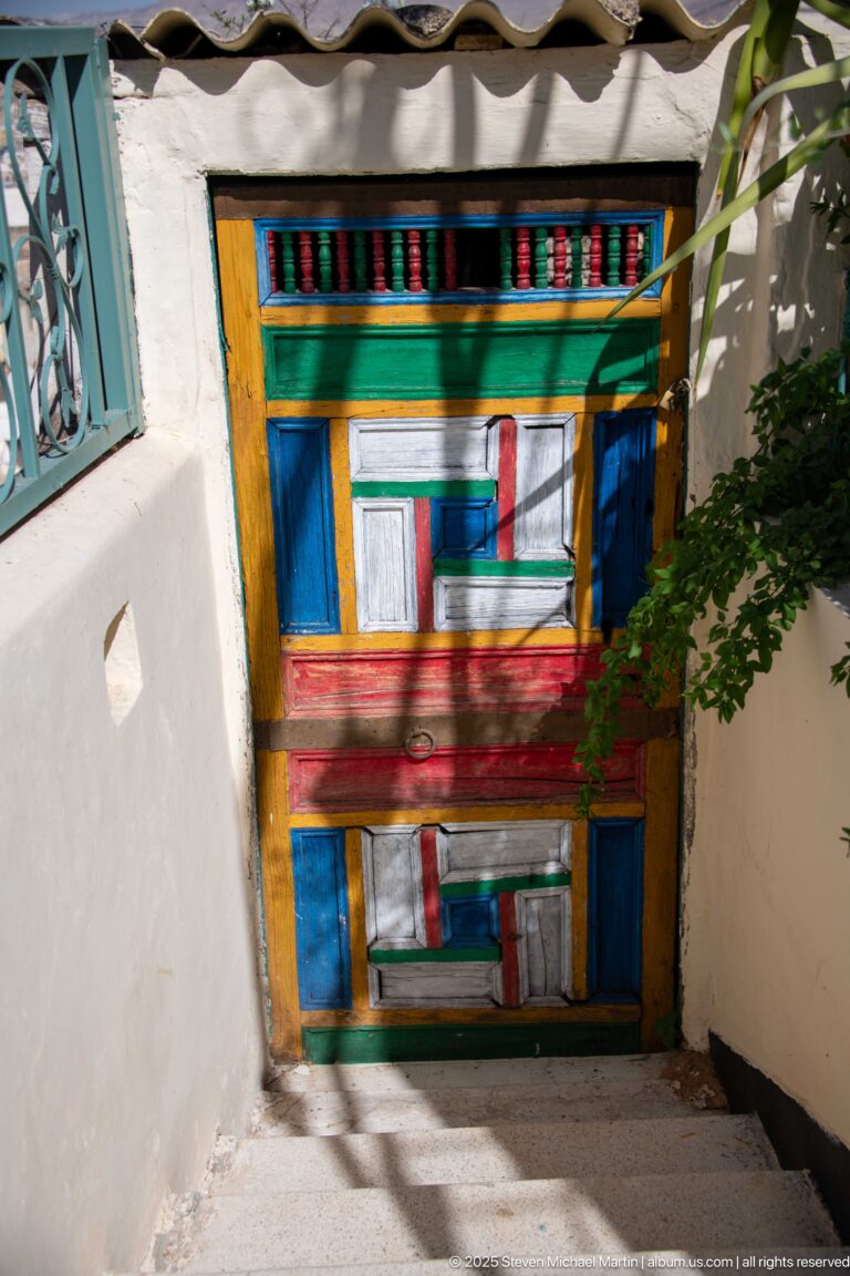 Bright multicolored wooden door with rectangular panels and patterned lintel in a sunlit narrow passage.