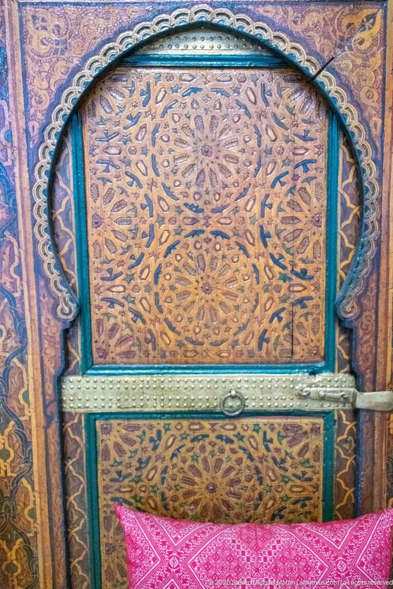 Close-up of an arched carved wooden panel with turquoise trim, dense vegetal patterning, metal brace, and pink cushion below.