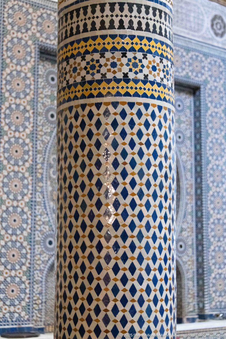 Close-up of a cylindrical column covered in blue, white, ochre, and dark geometric zellij, with tiled wall blurred behind.