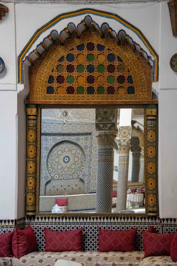 Large ornate mirror with colored-glass crest above red cushions, reflecting zellij walls, an arched niche, and columns.