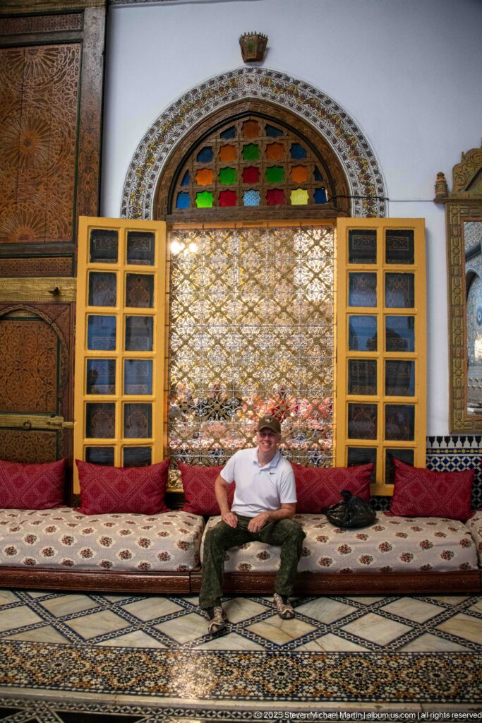 Seated visitor on a low patterned salon bench beneath a pierced screen and arched multicolored-glass window.
