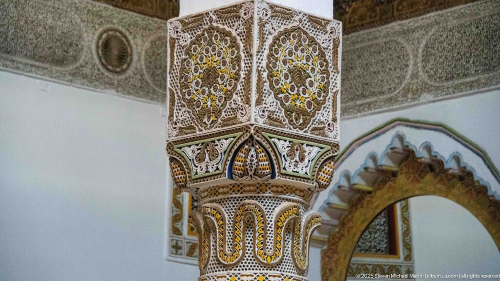 Close view of a richly carved square column capital with colored medallions and scalloped forms beside a cusped arch.