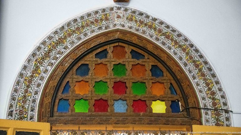 Arched window with multicolored glass panes and dark wooden tracery, surrounded by concentric floral and geometric ornament.