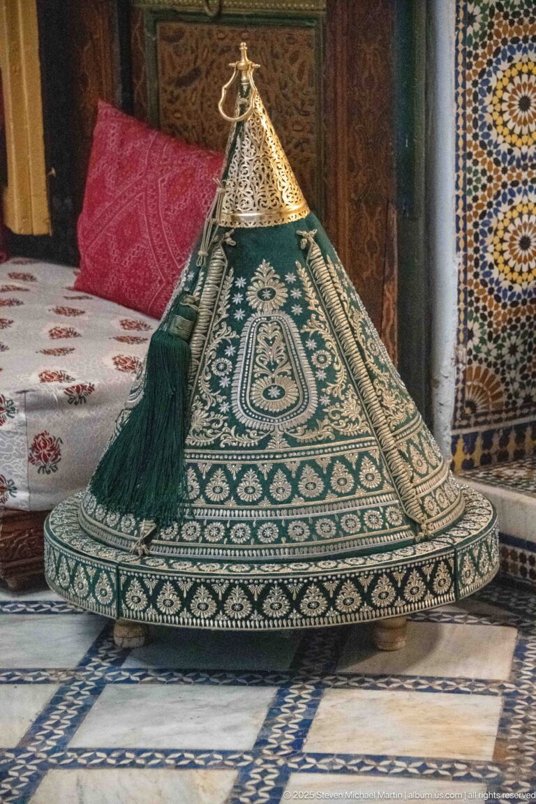Green conical textile cover with cream embroidery, patterned round base, long tassel, and metal finial.