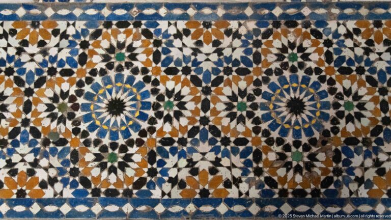 Close-up of zellij with two large blue-centered radial rosettes amid interlocking white, green, ochre, and dark geometric tiles.