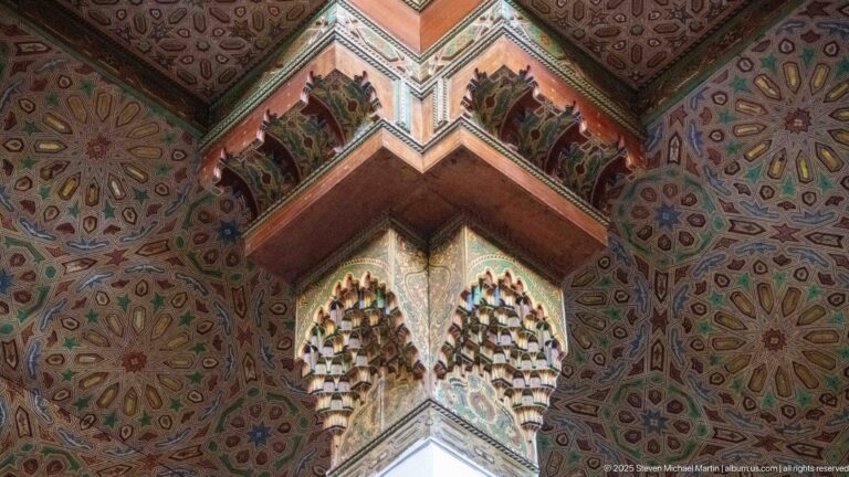 Upward corner view of a muqarnas-like capital beneath painted ceiling surfaces filled with multicolored rosettes.