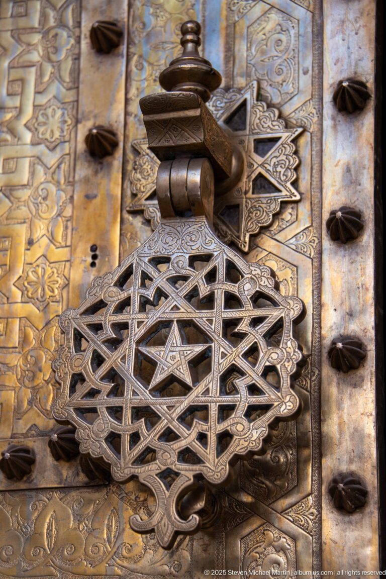 Close-up of an interlaced star-shaped metal door fitting mounted on a carved wooden door with dark bosses.