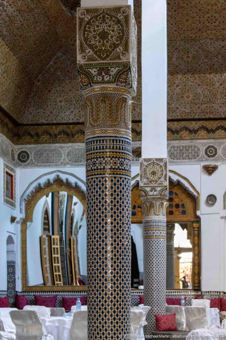 Two decorated columns, one clad in geometric zellij, dividing a palace hall with arches, salon seating, and white-covered tables.