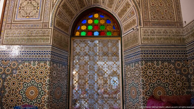 Arched window with red, green, blue, and amber glass above a pale pierced screen, surrounded by zellij and ornament.