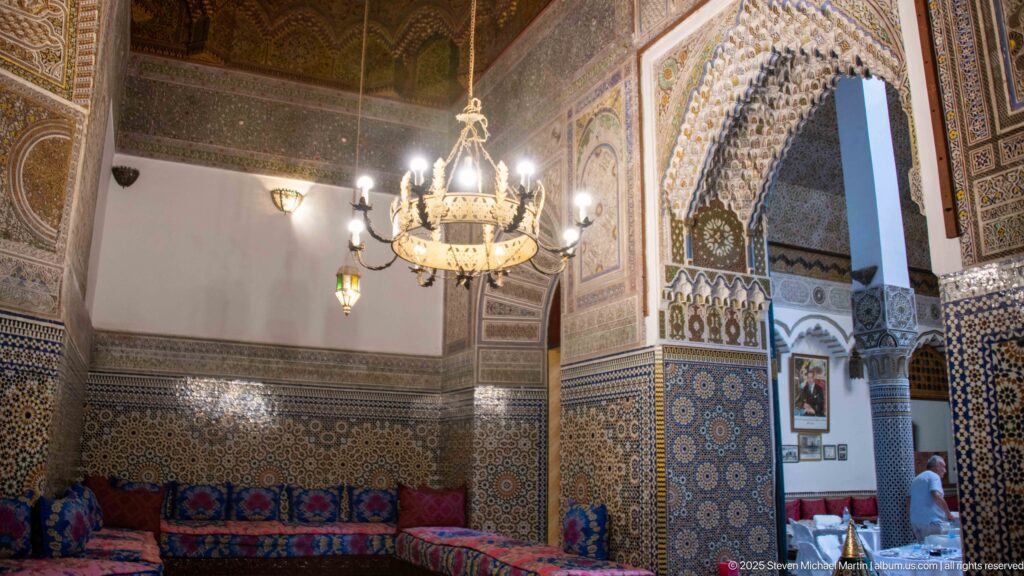 Palais Mnebhi salon with chandelier, low cushioned seating, zellij walls, and an ornate arch opening to another room.